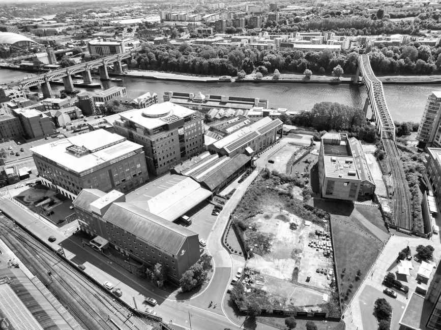 An aerial black-and-white photograph showing a cityscape with a river running through it, flanked by numerous buildings, roads, and green spaces. In the foreground, there are several large industrial